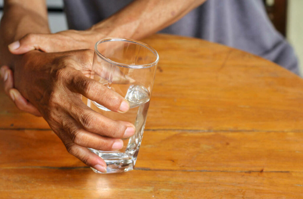 An old man is holding a glass of water in his right hand while his left hand is supporting and holding his right hand to control the trembling of his hand.