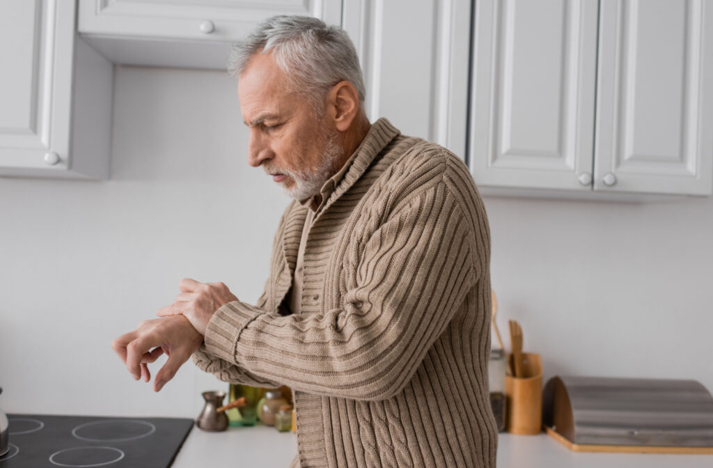 A senior man standing in his kitchen, holding onto his right wrist with his left hand to stop it from trembling.