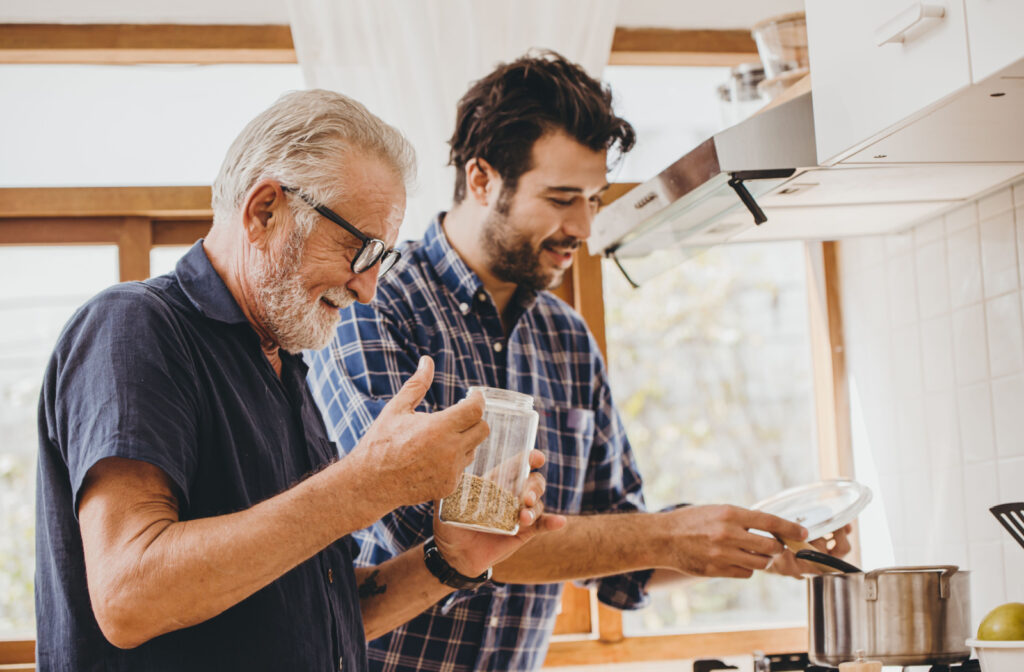 A son wearing a plaid shirt assisting his senior father in preparing oats for breakfast.