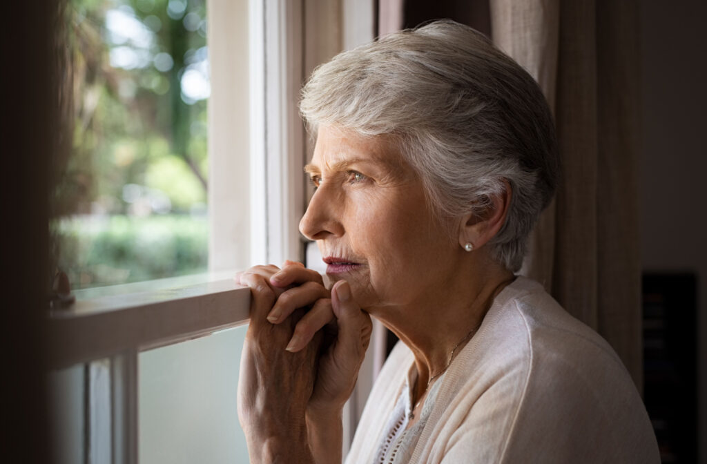 An older adult woman holding onto a window sill and looking out of the window with a serious expression.
