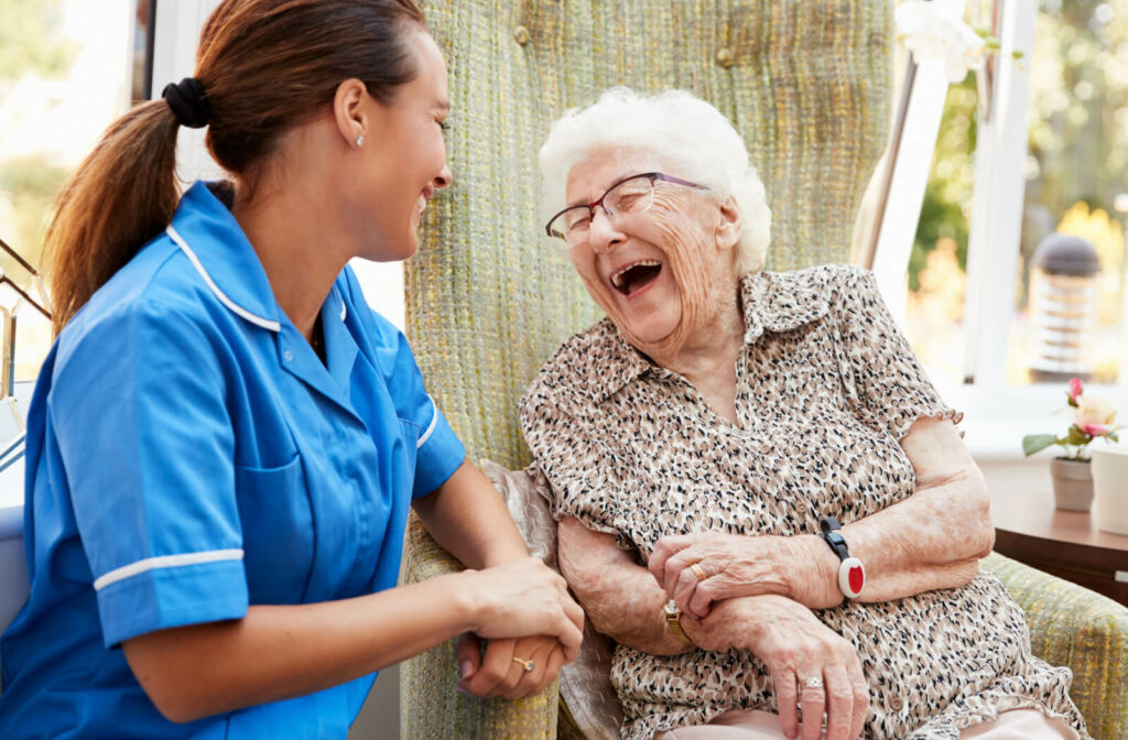 An older adult woman in a memory care  facility sitting on a chair smiling and having a conversation with a nurse.